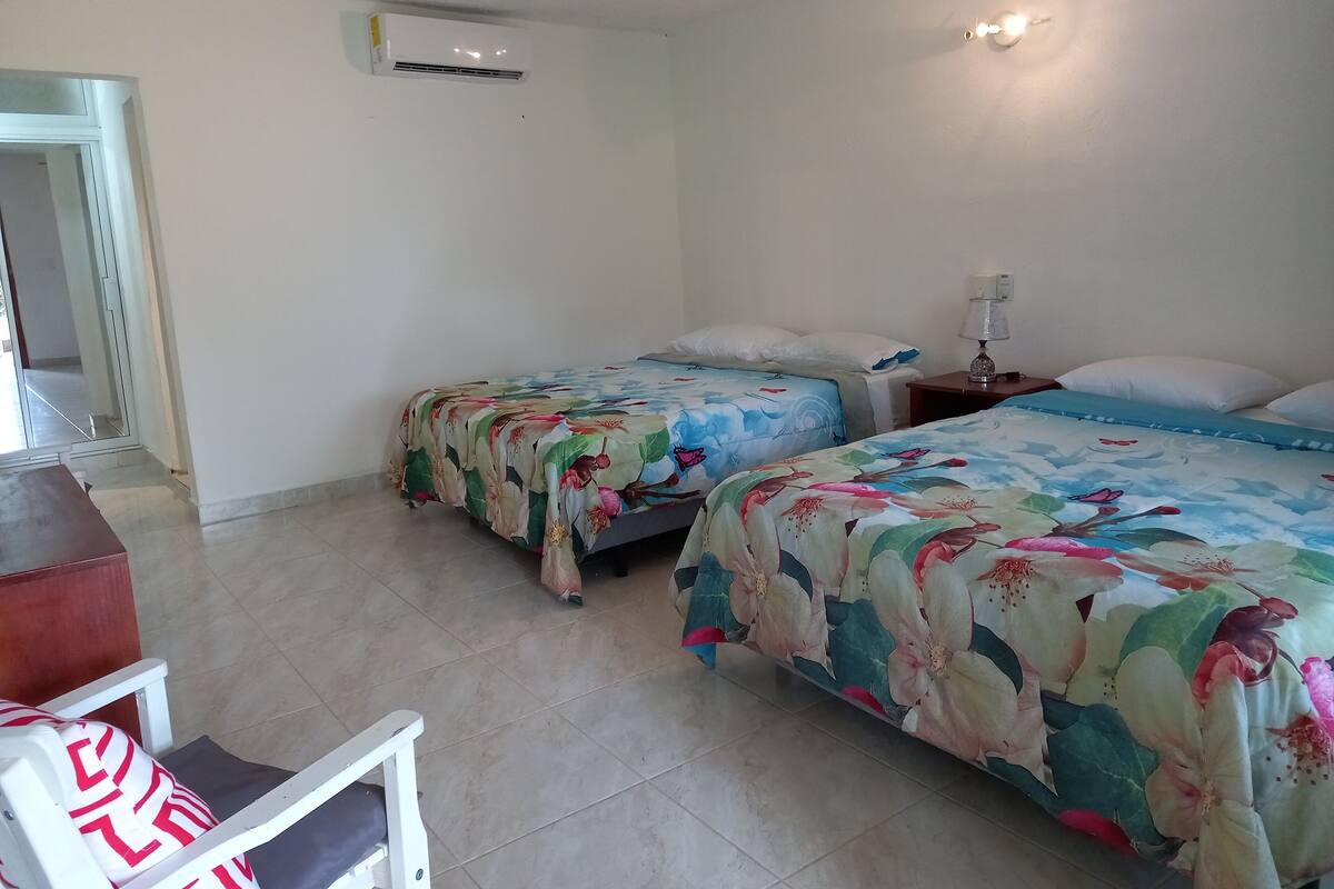 Een hotelkamer met twee bedden, een plafondventilator, een airconditioningapparaat, een nachtkastje met een lamp en een stoel met een gestreept kussen.
