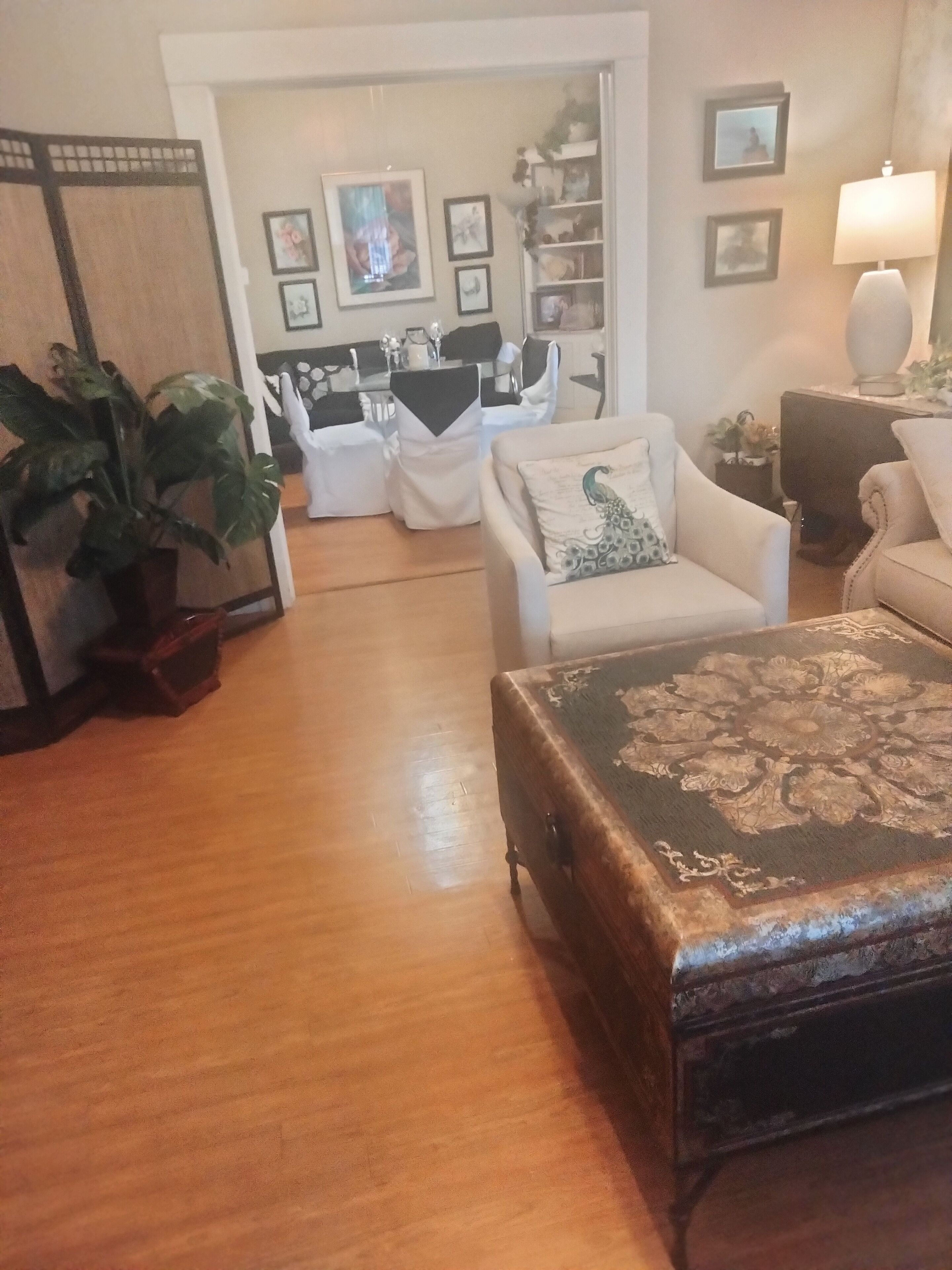 A living room with a wooden floor, a white sofa with a peacock pillow, a vintage trunk, and a dining area with white chairs and a table.