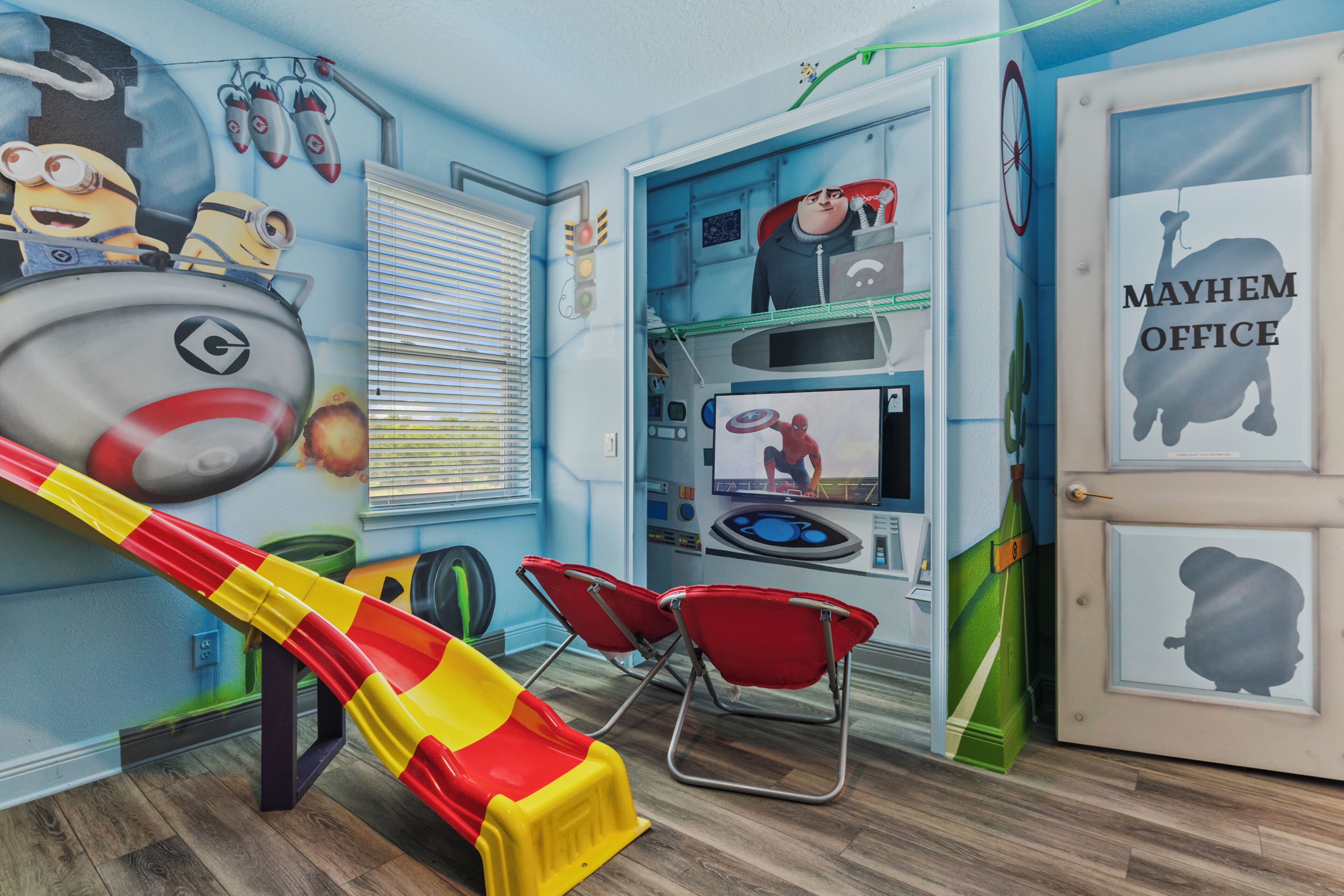 Legeområde for børn med en rutchebane, røde stole og en væg udsmykket med Minion-figurer og et skilt fra Mayhem Office.