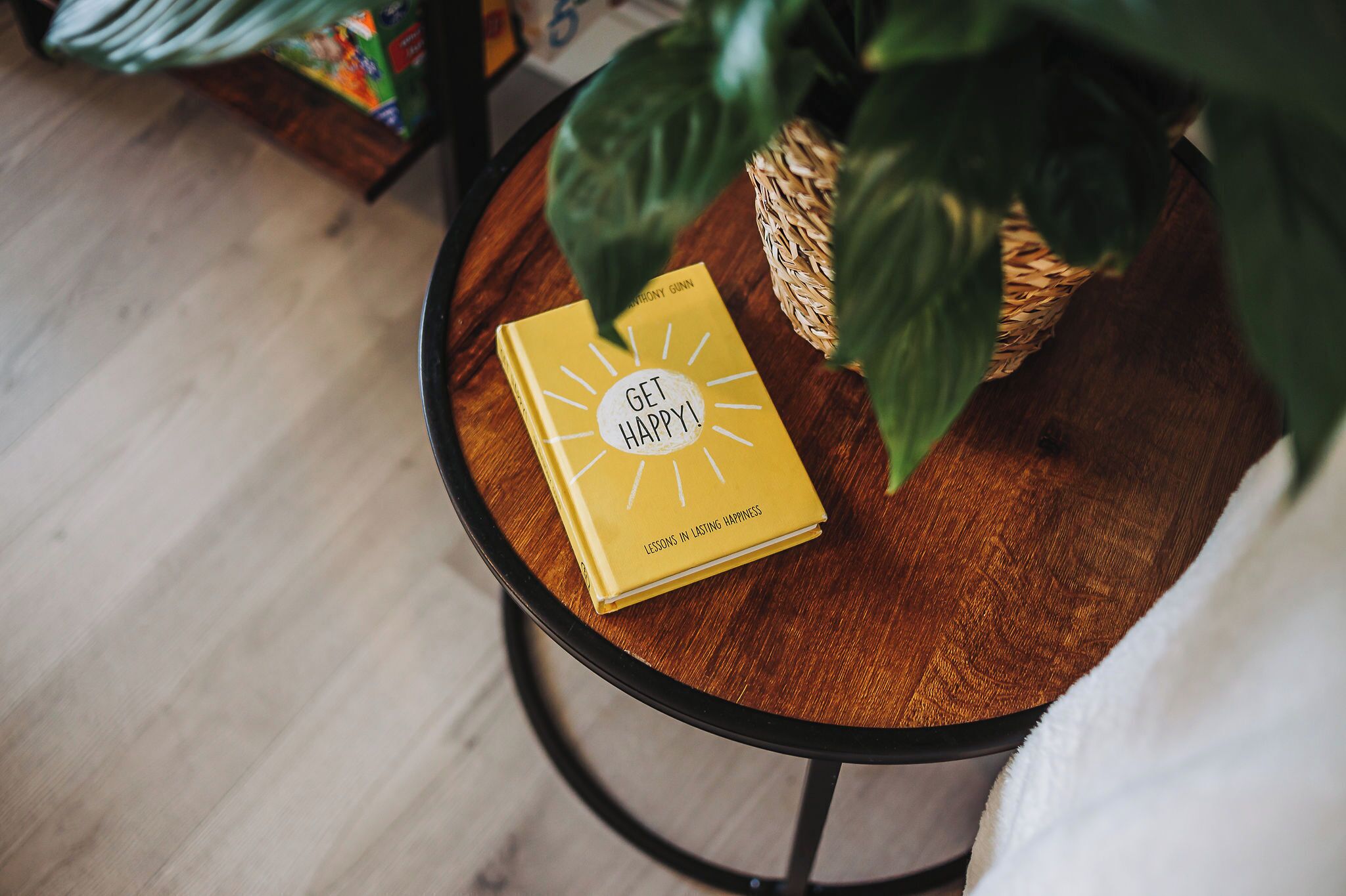 A book titled "Get Happy!" on a round wooden table with a woven basket and a green plant beside it.