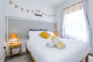 A neatly made bed with white bedding and a rolled towel, a bedside lamp, and a small table with a plant.
