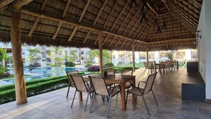 Un espace extérieur aménagé pour les repas, doté d’un toit de chaume et entouré d’une piscine et de verdure.