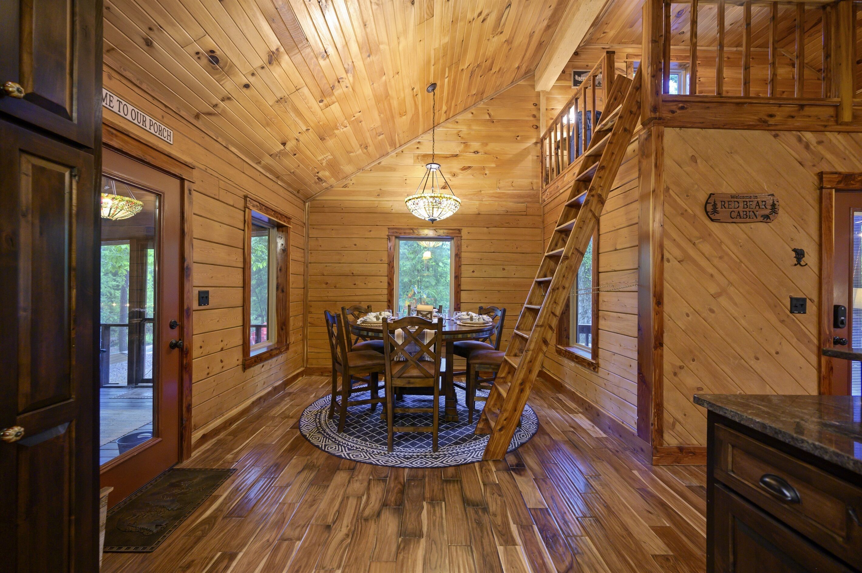 A wooden cabin interior with a dining table, chairs, a chandelier, and a staircase.