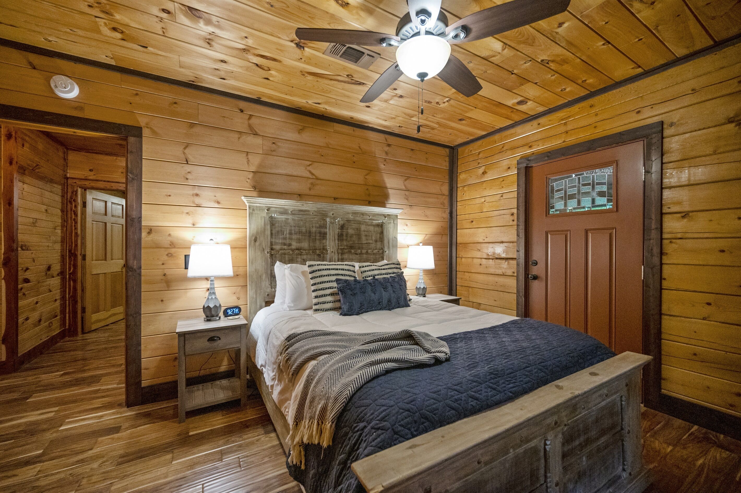 A wooden-paneled bedroom with a bed, nightstands, a ceiling fan, and a door with a window.