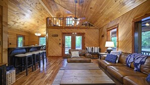 Ein Holzblockhaus-Interieur mit Schlafzimmer im oberen Bereich, Essbereich und Wohnzimmer, möbliert mit Ledersofas und einem Couchtisch.