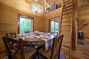 A wooden cabin dining room with a large table set for four, wooden chairs, a chandelier, and windows with a view of trees.