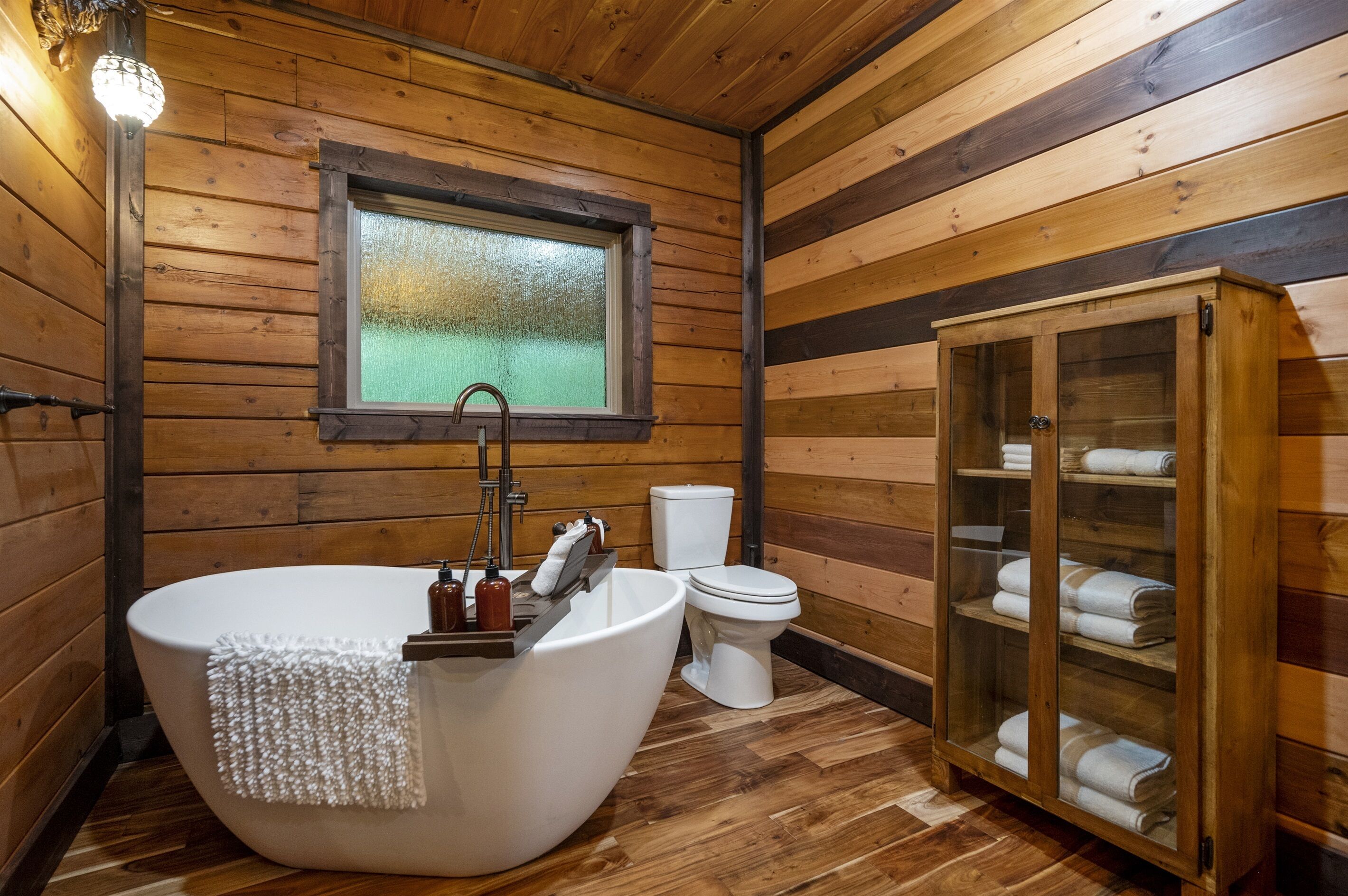 Une salle de bain rustique, avec des murs en bois, comprenant une baignoire sur pieds, des toilettes et un meuble en verre contenant des serviettes.