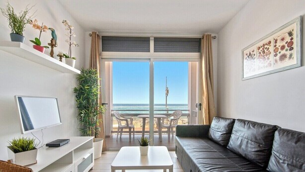 A living room with a TV, white coffee table, and a sofa. There are plants on a shelf and a balcony with a view of the beach and a tower.