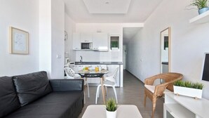 A modern living room with a black sofa, a dining table with chairs, a kitchen area with white cabinets and appliances, and a small balcony with plants.