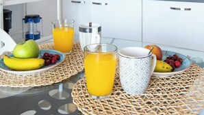 A modern kitchen with white cabinets, a microwave, a blender, and a sink. There is a clock on the wall and a table with fruit and a glass of orange juice.