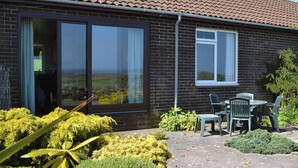 A house with a tiled roof, a large sliding glass door, and a small window. There is a patio with a table and chairs.