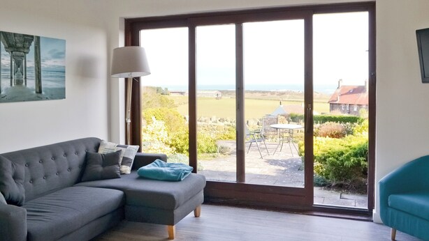 A living room with a grey sofa, a teal armchair, a floor lamp, and a large sliding glass door leading to a garden with a table and chairs.