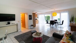 A modern living room with a grey sofa, a coffee table, and a flat-screen TV. There is a dining area with a table and chairs, and a kitchen in the background. - Apartment Oasis - Gemütliche Wohnung mit Garten (Mendig)