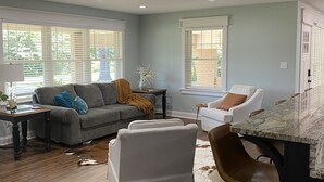 Una sala de estar moderna con un sofá gris, sillones blancos y una zona de cocina con encimera de mármol.