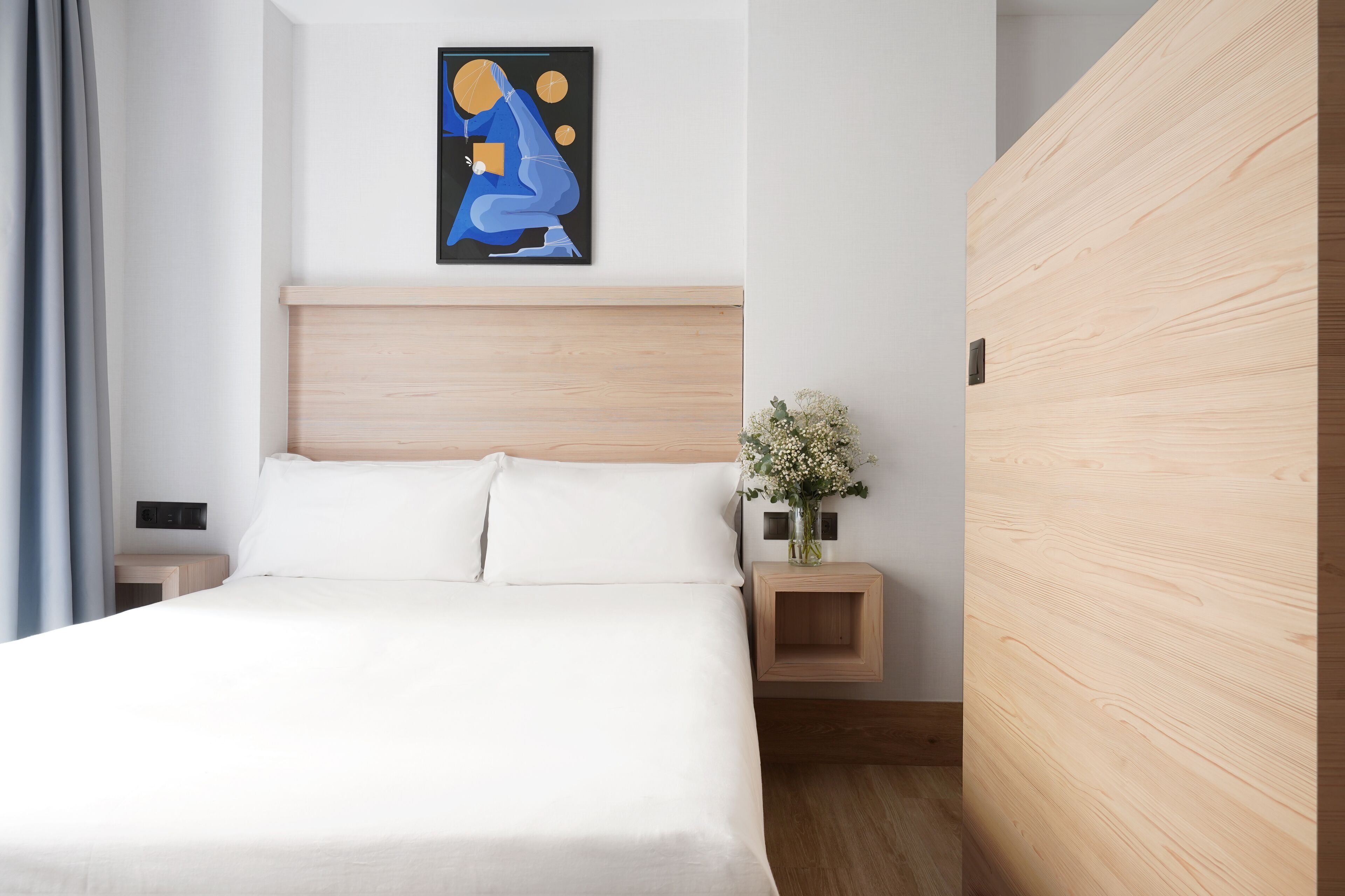Une chambre à coucher moderne avec une tête de lit en bois, du linge de lit blanc, un tableau abstrait accroché au mur et une table de chevet ornée d’un vase de fleurs.