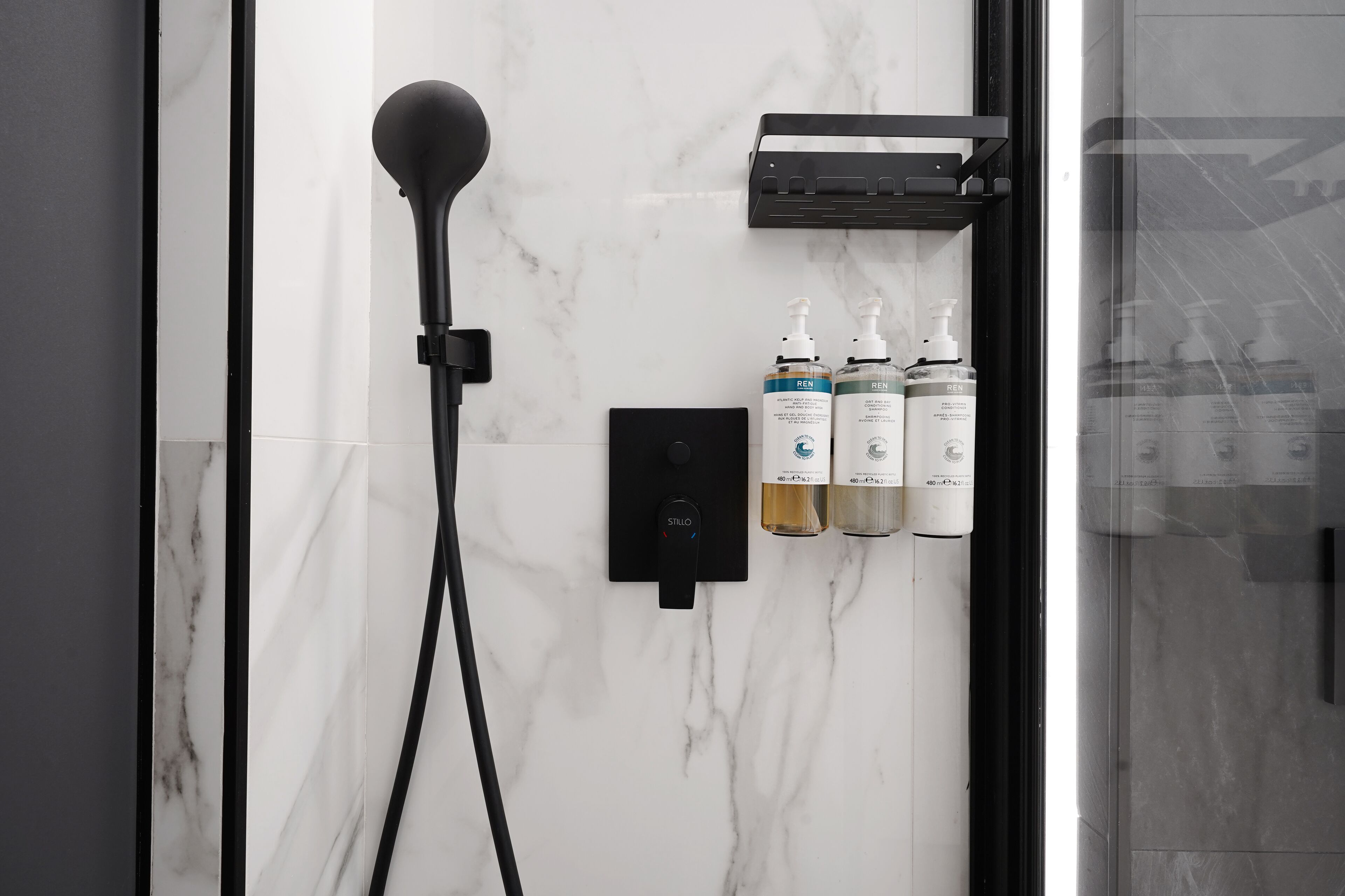 Une salle de bain moderne avec un mur en marbre, une pomme de douche noire et deux flacons de gel douche de la marque REN.