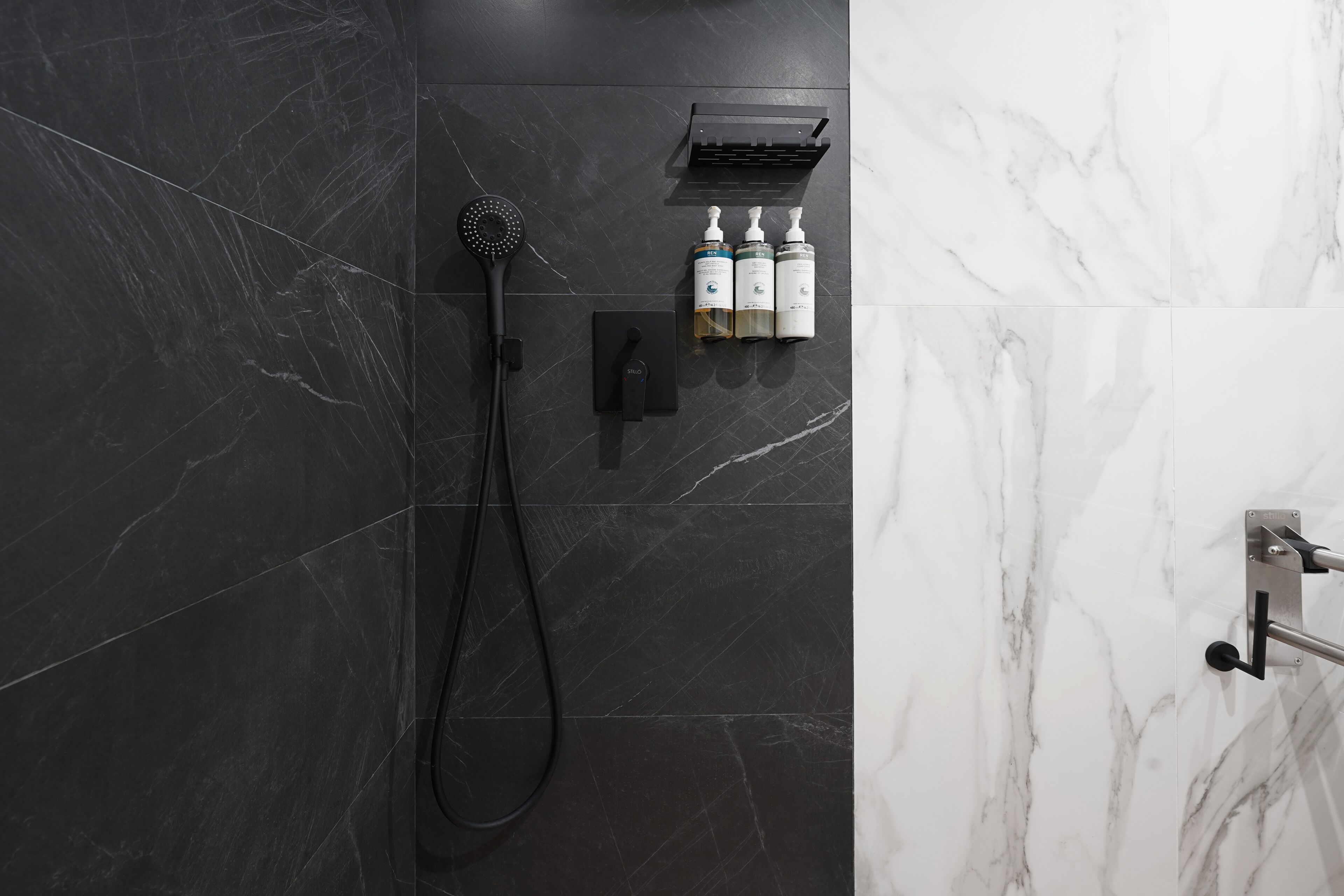 A modern bathroom with a black marble shower, a handheld showerhead, and two bottles of shampoo and conditioner on a shelf.