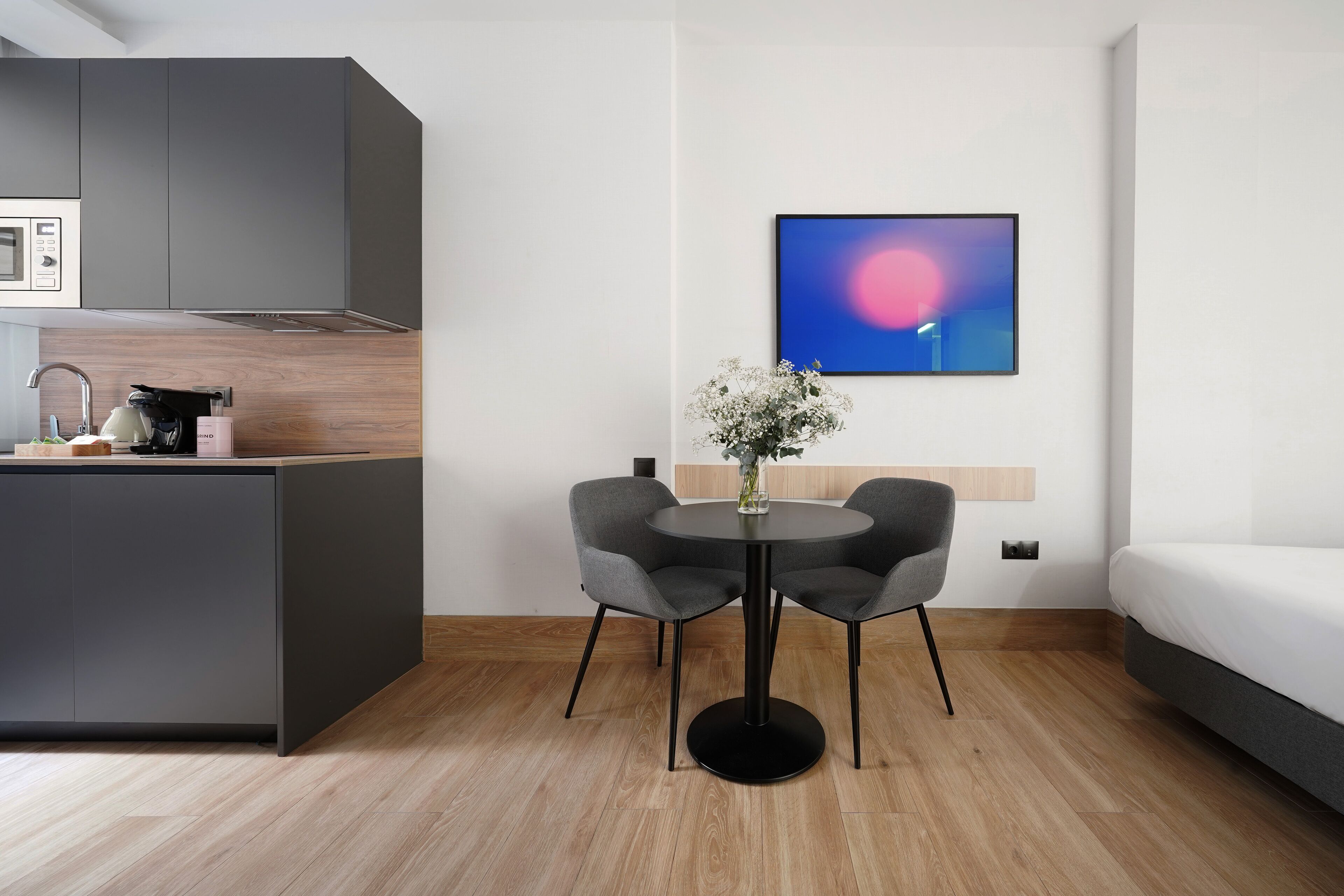 A modern room with a kitchen area, a round table with two chairs, a bed, and a wall-mounted artwork.