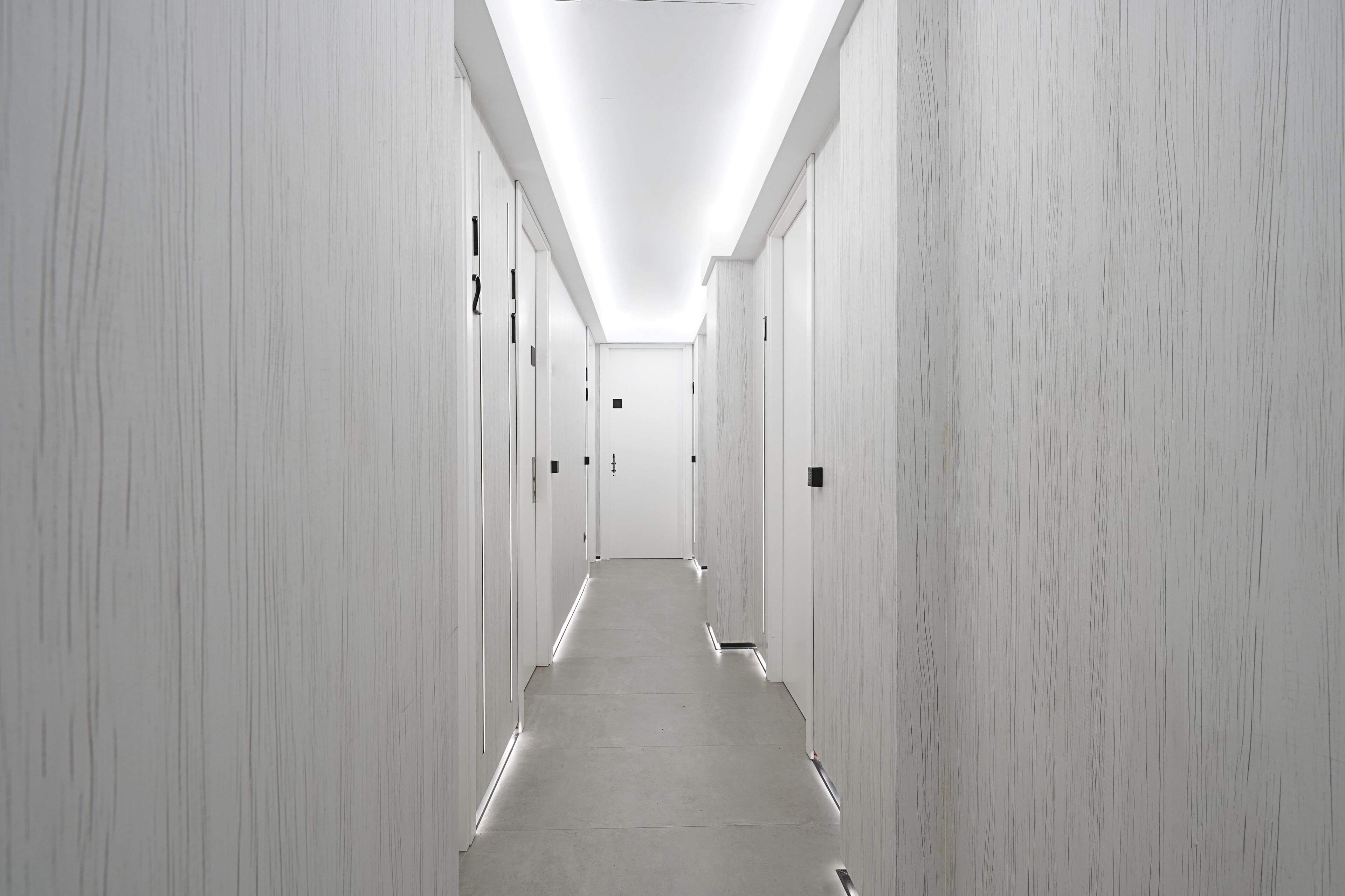 A long, narrow hallway with white walls and doors on both sides, leading to a door at the end.