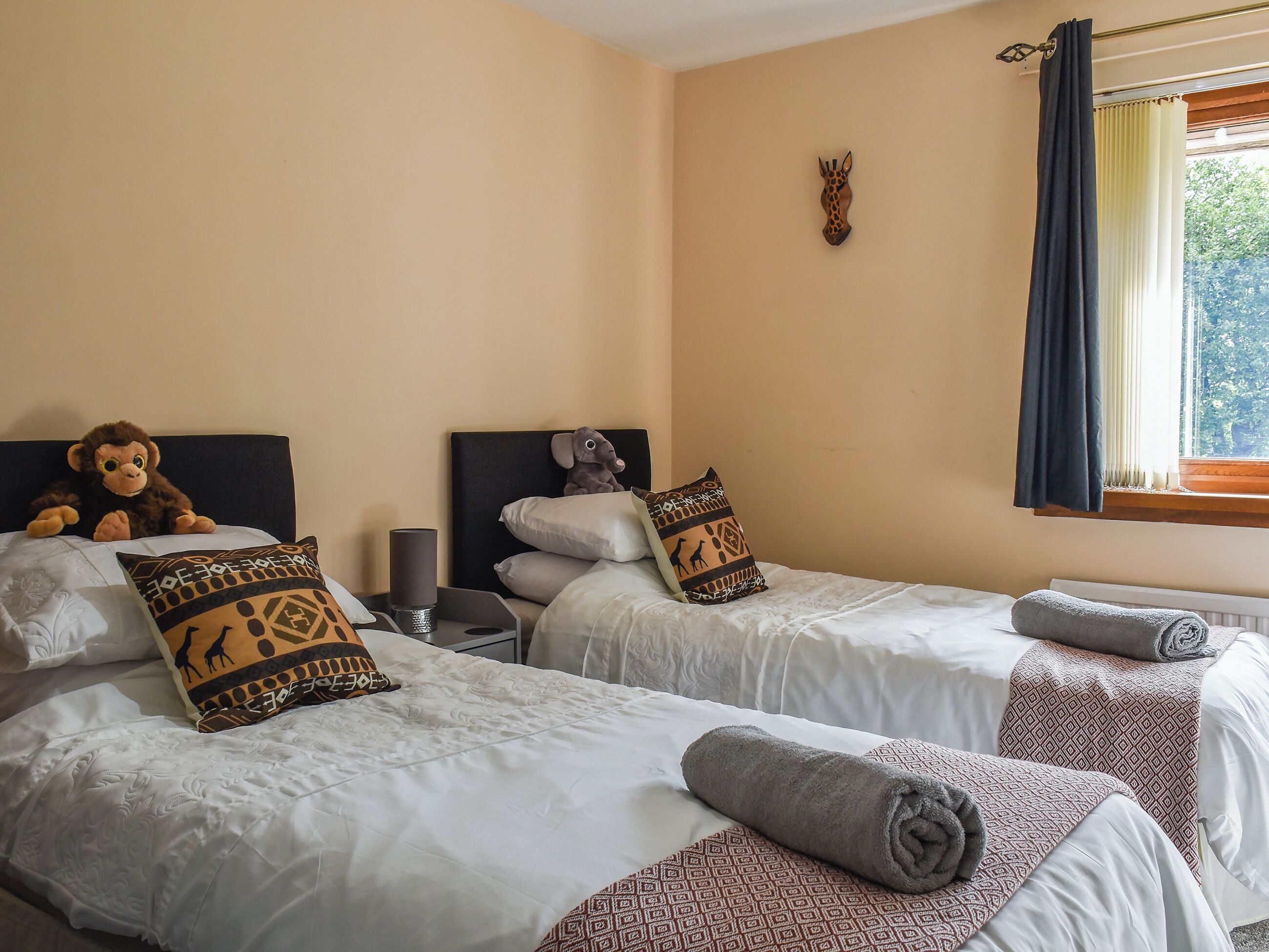 Habitación de hotel con dos camas, cada una con sábanas blancas y almohadas estampadas. Hay una mesita de noche con una lámpara, un adorno de pared y una ventana con cortinas.