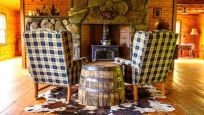 Un intérieur de chalet chaleureux avec une cheminée en pierre, deux fauteuils à carreaux, un tonneau en bois et un tapis en peau de vache.