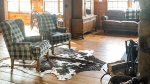 L’intérieur d’un chalet rustique, avec un plancher en bois, une grande fenêtre, un coin repas et un salon comprenant un canapé et des fauteuils.