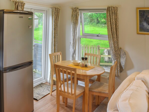 A compact kitchen with a refrigerator, wooden dining table, chairs, and a view of a garden through the window.