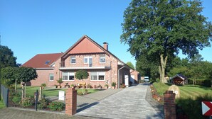 A two-story brick house with a red-tiled roof, a paved driveway, and a large tree in front. - Vacation apartment in a quiet, rural location (Großefehn)