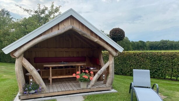 A wooden gazebo with a bench and a table, surrounded by a hedge and a lawn. - Vacation apartment in a quiet, rural location (Großefehn)