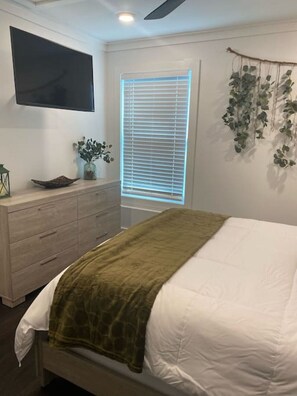 Ein Schlafzimmer mit einem Bett, einer Kommode, einem an der Wand befestigten Fernseher und einem Fenster mit Jalousien.