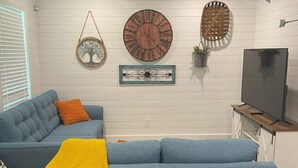 Un salon avec un canapé bleu, un ventilateur de plafond, une horloge murale et un téléviseur.