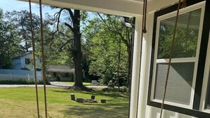 Une balançoire verte, installée sur un porche, avec un coussin à carreaux, donnant sur une cour verdoyante où se trouvent une maison et des arbres.