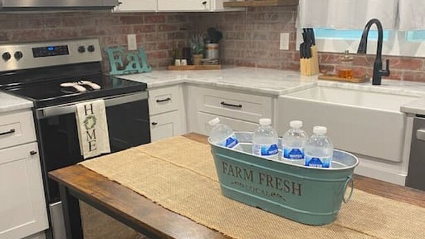 Une cuisine avec des armoires blanches, un four à micro-ondes en acier inoxydable, une cuisinière noire et un évier avec un robinet noir. On y trouve une table en bois surmontée d’un set de table en fibres tressées et un seau bleu portant l’inscription « PRODUITS FRAIS DE LA FERME », contenant des bouteilles d’eau.
