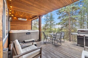 Relax on the deck with a hot tub, grill, and seating area, surrounded by a forested landscape.