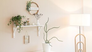 Una mesita de noche blanca con una planta en maceta, un estante de pared con objetos decorativos y una lámpara de pie.