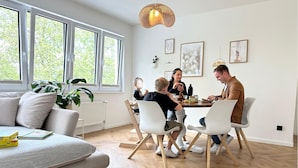 Una sala de estar moderna con una mesa de comedor, sillas blancas y un sofá. Hay ventanas amplias, una planta y obras de arte en las paredes.