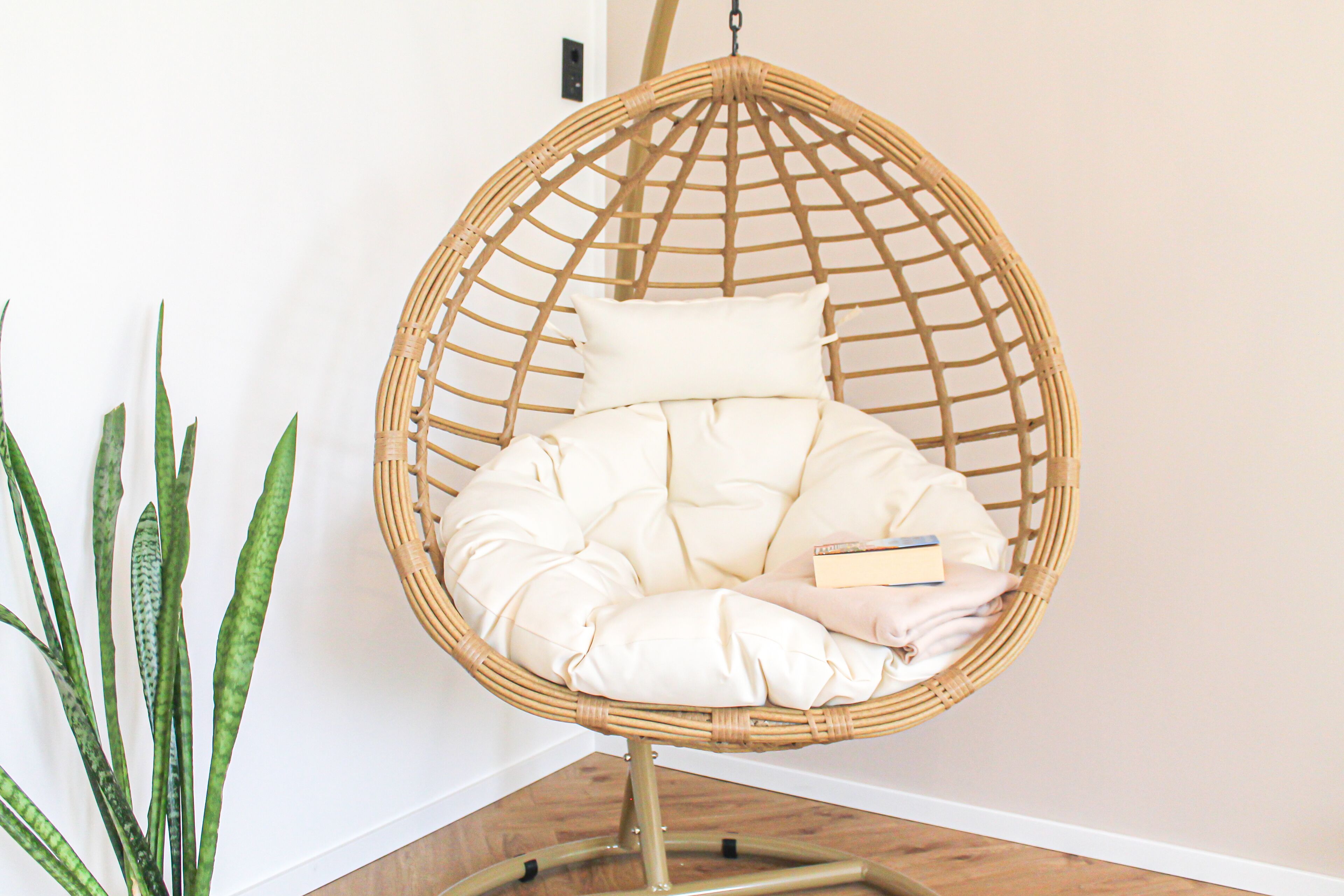 Una silla colgante de mimbre con asiento y respaldo acolchados, ubicada frente a una pared con una planta en maceta a su lado.