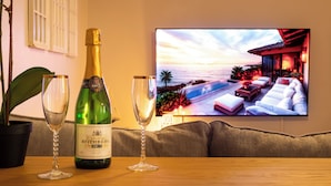 Una botella de champagne Brut Rothberg y dos copas sobre una mesa de madera, con un televisor mostrando una vista de atardecer de una piscina y un área de descanso.