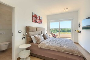 A bedroom with a large bed, a bedside table, a wall-mounted TV, and a window with a view of greenery and buildings.