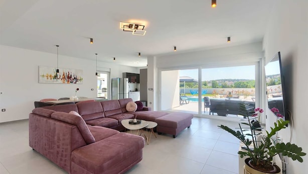 Een ruime woonkamer met een grote roze hoekbank, een klein rond bijzettafeltje en een potplant. Er is een glazen schuifdeur die leidt naar een buitenruimte met een zwembad en stoelen.