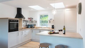 A modern kitchen with a built-in oven, a microwave, and a coffee maker. There are two stools at the island and a window with a view of greenery.