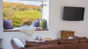 A room with a large window offering a view of a green landscape, a brown leather sofa, a wooden side table with a teapot and cups, and a flat-screen TV mounted on the wall.