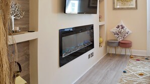 Un salon moderne doté d’une cheminée, d’un téléviseur à écran plat fixé au mur et d’un coin salon confortable avec une petite table ronde et un pouf.