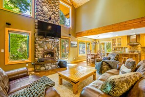 A spacious living room with a stone fireplace, leather furniture, and large windows offering views of the outdoors.