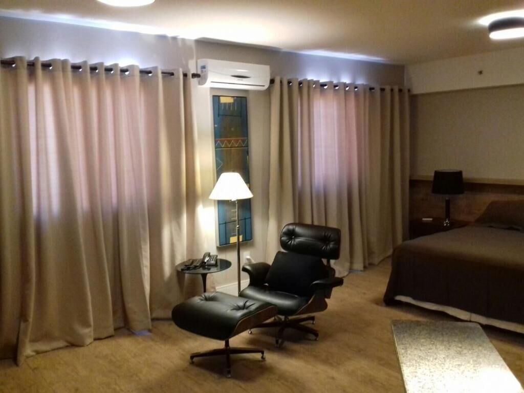 A hotel room with a bed, a black leather chair, a small table with a lamp, and beige curtains.