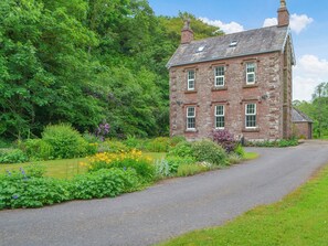 Une maison en pierre avec une allée pavée, entourée d’une végétation luxuriante et d’une grande variété de fleurs.