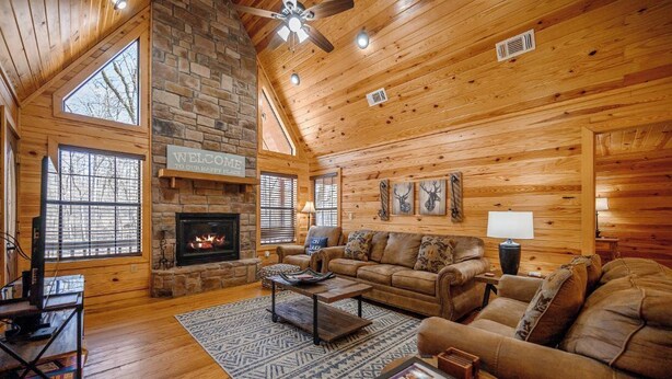A cozy log cabin interior with a fireplace, wooden walls, a leather sofa, and a large window. - Feisty Goat - Cozy Cabin, Sleeps 6, Hot Tub Overlooking Creek. (Broken Bow)