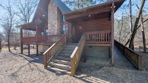 A log cabin with a stone chimney, wooden deck, and a staircase leading to a porch. - Feisty Goat - Cozy Cabin, Sleeps 6, Hot Tub Overlooking Creek. (Broken Bow)