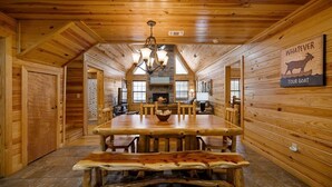 A wooden cabin interior with a large dining table, a fireplace, and a 'Welcome' sign. - Feisty Goat - Cozy Cabin, Sleeps 6, Hot Tub Overlooking Creek. (Broken Bow)