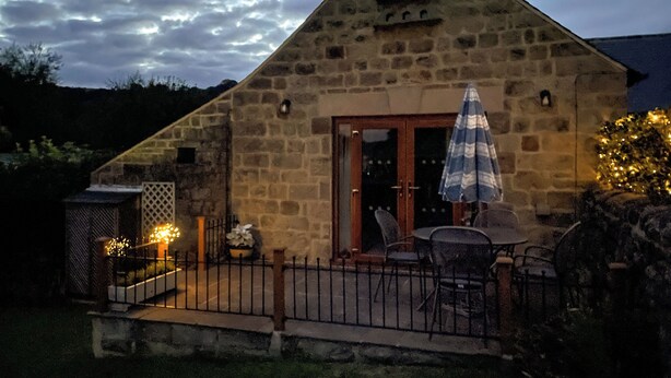 A stone building with a triangular roof, a wooden door, and a small patio with a table and chairs.